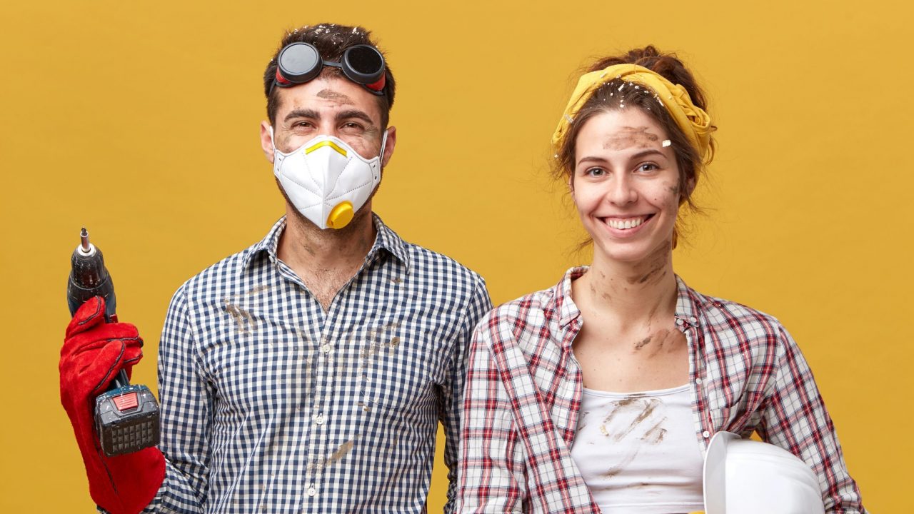 Studio portrait of cheerful male wearing protective mask, goggles and gloves holding drill fixing something at house and his wife who is helping him with construction holding hardhat. Service workers